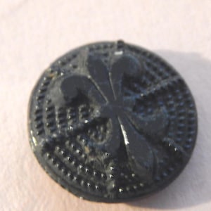 May include: A black, round button with a fleur-de-lis design in the center. The button has a textured surface and is set against a light pink background. The button's design is raised, creating a three-dimensional effect.