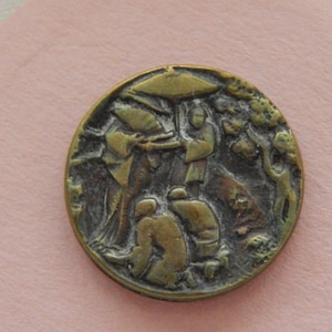 May include: A circular brass button featuring a detailed relief scene of figures under an umbrella. The metallic surface has a dark, aged patina with raised golden highlights depicting people in traditional attire amidst trees and foliage.