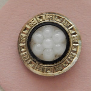 May include: A circular button featuring a central cluster of seven white, rounded beads arranged in a flower pattern. The center is framed by a thin black ring, surrounded by a metallic gold-toned border with a repeating geometric pattern.