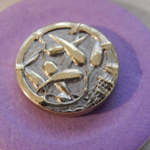May include: A gold and silver button with a design of stylized olive branches. The button is on a purple background.