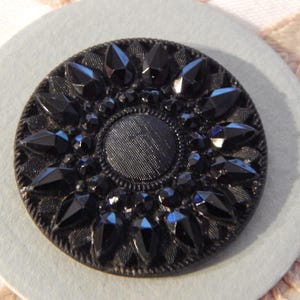 Könnte beinhalten: Ein schwarzer Knopf mit einem strukturierten Mittelkreis, der von einem Kranz aus schwarzen, facettierten, edelsteinartigen Verzierungen umgeben ist. Der Knopf befindet sich auf einer hellgrauen, kreisförmigen Basis, mit einem weißen und pfirsichfarbenen Blumendesign im Hintergrund.