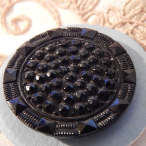 Könnte beinhalten: Ein schwarzer Glasknopf mit einem erhabenen, strukturierten Design. Der Knopf ist rund mit einer erhabenen, strukturierten Mitte und einem schwarzen, strukturierten Rand.