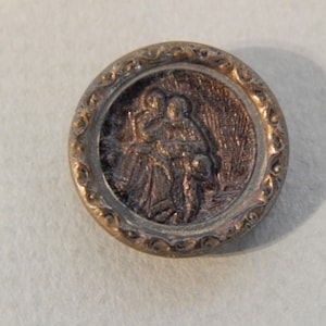 May include: A gold-toned metal button with an ornate design around the edge. The center of the button features a raised image of two people standing together.