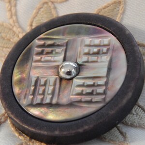 Peut inclure: Un bouton rond avec un bord marron foncé et un centre en nacre. Le centre présente un motif géométrique et un petit clou argenté. Idéal pour la couture ou les travaux manuels.
