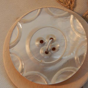 May include: A large, round button with a mother-of-pearl finish, featuring a smaller, decorative button on top. The button has four holes and a light-colored wooden base. The top button has a circular design.