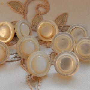 Puede incluir: Colección de diez botones redondos vintage. Cada botón presenta un disco central nacarado rodeado por un borde decorativo y estriado. Los botones, de color blanquecino, están dispuestos sobre una tela clara, resaltando su diseño.