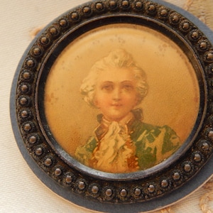May include: A vintage round brooch with a portrait of a person in a green jacket and white cravat. The portrait is set within a dark, ornate frame with a beaded edge, mounted on a blue backing.