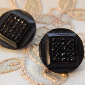 May include: Two black buttons with a square of small, dark gems set in the centre. The buttons are round with a square inset. The buttons are on a white surface with a floral design.
