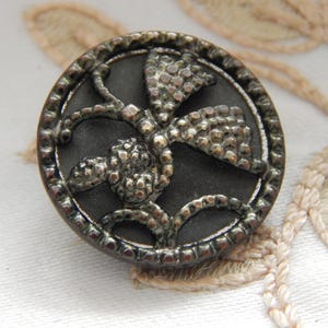 May include: A round, antique button with a dark gray center and a detailed, raised floral design in a silver-tone metal. The button's edge is also silver, with a beaded texture. The button is likely for sewing or crafts.
