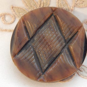 Puede incluir: Un botón redondo marrón con un diseño geométrico. El botón presenta una forma de diamante central con un patrón entrecruzado, rodeado de formas angulares. El botón probablemente está hecho de un material duro.