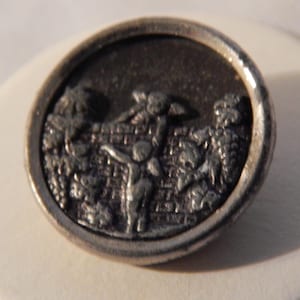May include: A silver-toned metal button with a raised design of people climbing a wall. The button has a textured surface and a circular shape.