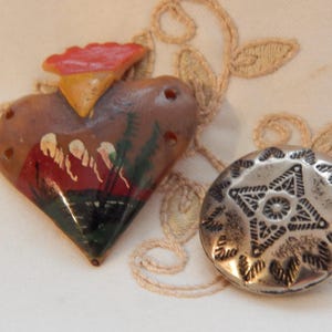 May include: Two vintage decorative buttons. One is heart-shaped, painted with a landscape scene in red, green, and brown. The other is a silver-colored, round button with a star design. Both are on a cream-colored surface.