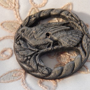 May include: A black, wooden button with a detailed carving of a bird perched on a branch. The button has two holes for sewing.