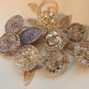 May include: A collection of decorative buttons in various shapes and sizes. The buttons are embellished with small, sparkling crystals in shades of silver, gray, and clear. Some buttons are oval, while others are round. The buttons are arranged on a cream-colored fabric.
