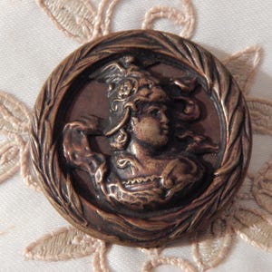 May include: A bronze button featuring a detailed image of a woman wearing a helmet and a flowing robe. The button is surrounded by a wreath of leaves.