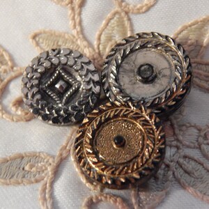 May include: Three antique buttons with intricate designs. One is silver with a diamond pattern, another is silver with a circular design, and the third is gold with a central black gem. They rest on a cream-colored, embroidered fabric.