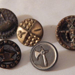May include: Five antique metal buttons with different designs. The buttons are round and have a variety of finishes, including gold, silver, and copper. The designs include a crescent moon, a shield, a stylized axe, a blacksmith's anvil, and a stylized figure.