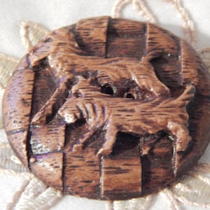May include: A round, brown button with a carved design of two dogs. The button has a textured, wood-like appearance and a striped background. The dogs are in a running or playful pose.