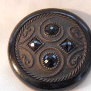 May include: A black button with a decorative design featuring four black glass stones and a raised, intricate pattern.