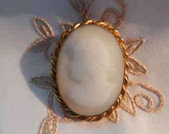 May include: A white cameo brooch with a gold-toned rope-like border. The cameo features a profile of a person's face.