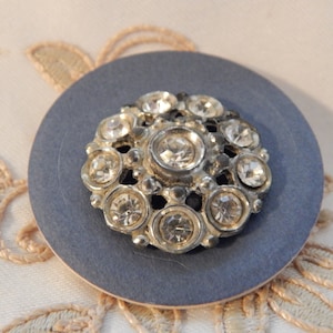 May include: A silver button with a circular design featuring a cluster of clear rhinestones.