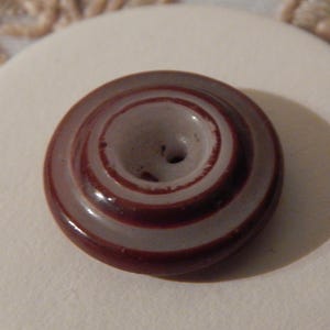 May include: A close-up of a vintage button with a spiral design. The button is a dark reddish-brown color with lighter, almost white, concentric rings. It has a central hole for sewing and sits on a white surface.