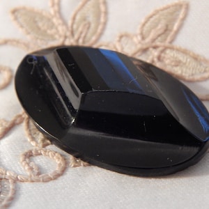 May include: A black, oval-shaped, glass button with a raised, geometric design. The button has a smooth, glossy finish.