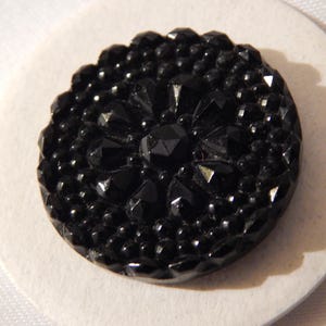May include: A black glass button with a raised, starburst design. The button is round and has a smooth, shiny finish.