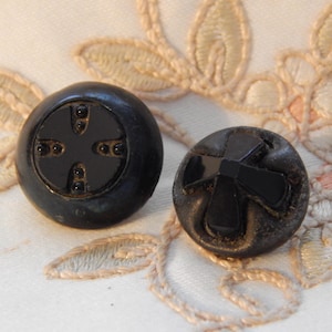 May include: Two round black buttons with a glossy finish. The left button features a recessed cross design with four small dots, while the right button displays a raised, faceted cross shape centered on its surface. Both items are circular.