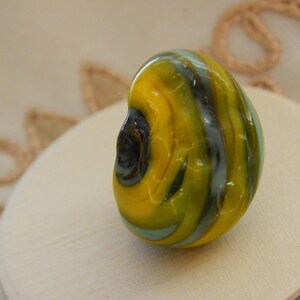 May include: A yellow, green, and blue glass bead with swirling patterns. The bead is smooth and shiny.