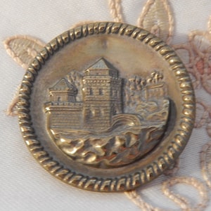 May include: A round, antique brass button with a detailed relief of a castle. The button has a rope-like border and the castle features towers, walls, and a landscape. The button is likely for sewing or crafts.