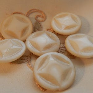 May include: Six off-white, round buttons with a starburst design. The buttons are arranged on a cream-colored surface with a subtle embroidered pattern. These vintage buttons are ideal for sewing and craft projects.