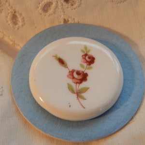 Può includere: Un piatto ovale in ceramica bianca con un bordo blu, con un delicato disegno floreale di rose rosa e steli marroni su sfondo bianco.