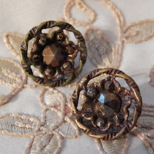 以下が含まれることがあります： 装飾的な華やかなデザインのアンティークな真鍮製のボタンが2つ。それぞれのボタンには、小さなファセットカットの石で囲まれた、中央にファセットカットされた虹色の石が特徴です。