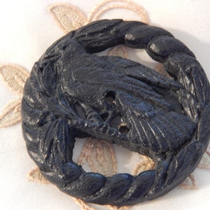 May include: A black button with a carved design featuring a raven perched on a branch within a circular wreath. The button has three holes for sewing and is set against a white background with a floral design.