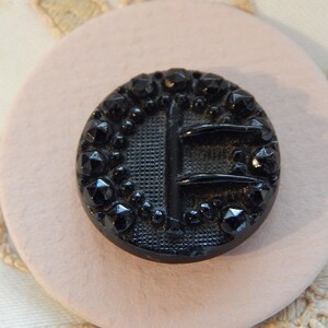 May include: A black, round button with a textured surface and a decorative design. The button features a central vertical bar and two horizontal bars, surrounded by a beaded border with faceted accents. The button is set on a light pink surface.