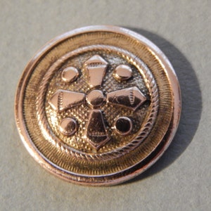 May include: A gold-toned button with a raised, intricate design featuring a cross shape with four smaller circles around it. The button has a silver-toned rim.