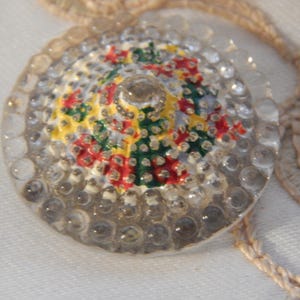 May include: A clear, round button with a domed center. The center features a painted floral design in red, green, and yellow. The button is surrounded by small, clear, bubble-like embellishments. A vintage sewing notion.