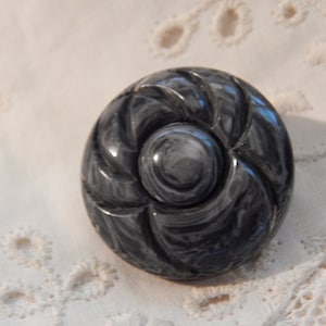 May include: A round, dark gray button with a swirling, marbled pattern. The button has a raised, central design and a carved, spiral-like pattern around the edges. The button is likely made of a hard material.