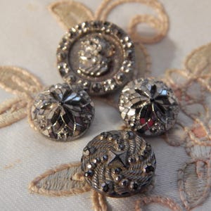 May include: An assortment of vintage silver buttons with intricate designs. One button features a classical profile, while others have faceted or textured surfaces. The buttons are arranged on a cream-colored fabric with delicate floral embroidery.