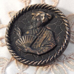 May include: A vintage metal button featuring a raised image of a woman in a traditional Japanese kimono holding a fan. The button is oval-shaped with a textured, rope-like border and a black background.