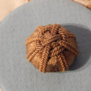 May include: A round, brown, woven button featuring a complex, interlaced cord pattern across its surface. The button is centered on a smooth, circular, light blue base, showcasing a detailed, textured knot design with a consistent, earthy brown tone.