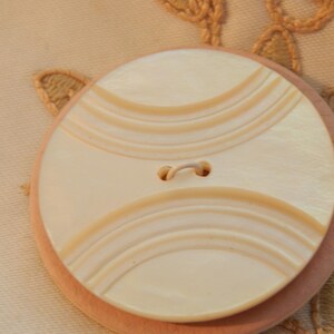 May include: A large, round button with a cream-colored face and a series of curved, light brown lines. The button has two holes and is set on a light pink base. The background features a cream-colored fabric with embroidered details.