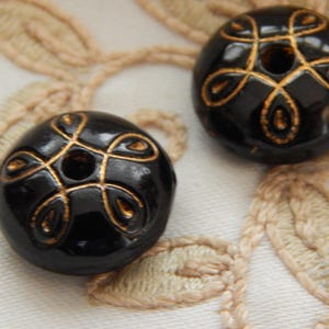 May include: Two black, round beads with a glossy finish. Each bead features a gold-colored, ornate design. The beads are resting on a cream-colored, embroidered fabric, showcasing the intricate details of the beadwork.
