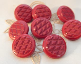 May include: A collection of eight round, red buttons with a geometric, diamond-shaped pattern. The buttons are glossy and appear to be made of a hard material. They are arranged on a light-colored surface.