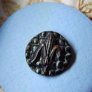 May include: A detailed close-up of a dark, antique button with a fleur-de-lis design. The button is round, with a textured, grid-like background. The fleur-de-lis is raised and has copper-colored accents. The button rests on a light blue circular surface.