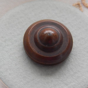 May include: A circular, dark brown button with a smooth, polished finish. The button features a raised, dome-shaped centre surrounded by two concentric, recessed rings that create a tiered, stepped profile leading to the outer edge.