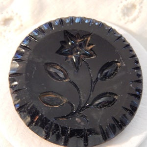 May include: A black glass button with a raised, carved design of a flower with five petals and leaves. The button has a scalloped edge.