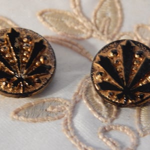 May include: Two round buttons with a black leaf design on a gold background. The buttons have a raised, textured surface and are set against a white background with a floral lace design.