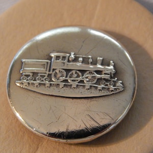 May include: A silver-toned button with a raised design of a train engine. The button is round and has a smooth surface.